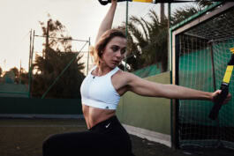 A person exercises with TRX bands on an outdoor court at sunset, surrounded by fencing and trees. 
