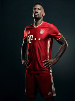 A person in a red FC Bayern Munich jersey, standing confidently against a dark background, hands on hips, showcasing tattoos on arms. 