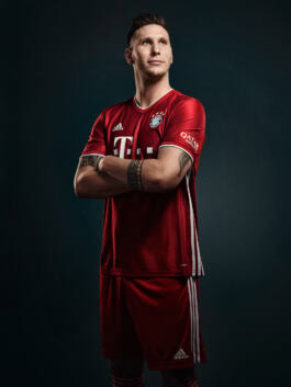 A person in a red Adidas sports uniform, arms crossed, poses confidently. The jersey features logos and stars, suggesting a sports team affiliation. 