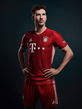 A person stands confidently in a red FC Bayern Munich soccer jersey, hands on hips, against a dark background. 