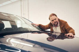 A person examines a car's shiny surface indoors, with a reflection visible on the hood. The background features a white brick wall. 