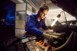 A person in a blue jumpsuit inspects a car engine with a work light in a garage setting. 