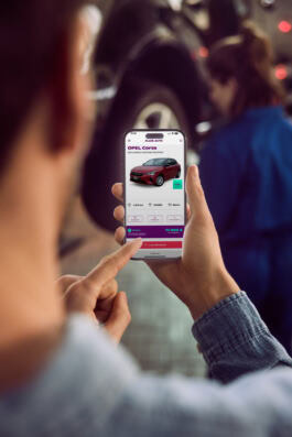 A person uses a smartphone with a car buying app, featuring an Opel Corsa, in a mechanic workshop setting. 