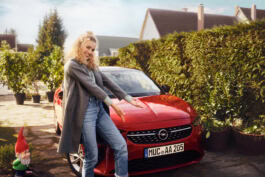 A person stands smiling beside a red car in a garden, with a small garden gnome nearby. 