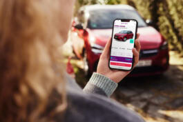 A person holds a smartphone displaying a car sale app, nearby is a red car, the background is blurred with greenery. 