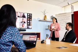 A person presents to others in a modern office, with virtual participants on a screen and a Henkel-branded laptop visible. 
