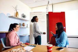 Three people collaborate in a modern office. One stands by a whiteboard. Shelves with decor and a globe are in the background. 