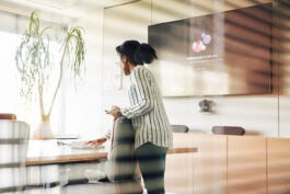 A person in a striped shirt stands in a modern office, using a device. A digital screen and large plant are present. 