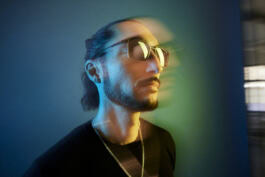 A person wearing sunglasses with a double exposure effect, creating a blurred movement illusion against a gradient blue and green background. 