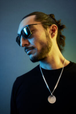 A person with sunglasses, wearing a necklace, poses against a gradient blue and green background, showing a profile with tied-back hair. 